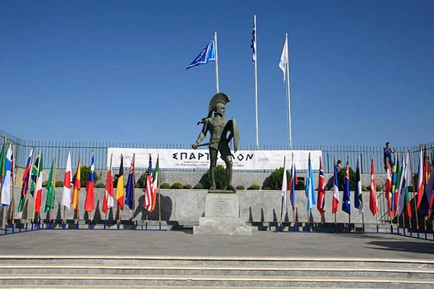 Llegada del Spartathlon en Esparta: Estatua de bronce del Rey Leónidas en su pedestal, rodeada de las banderas de los países participantes y la pancarta oficial en el fondo.